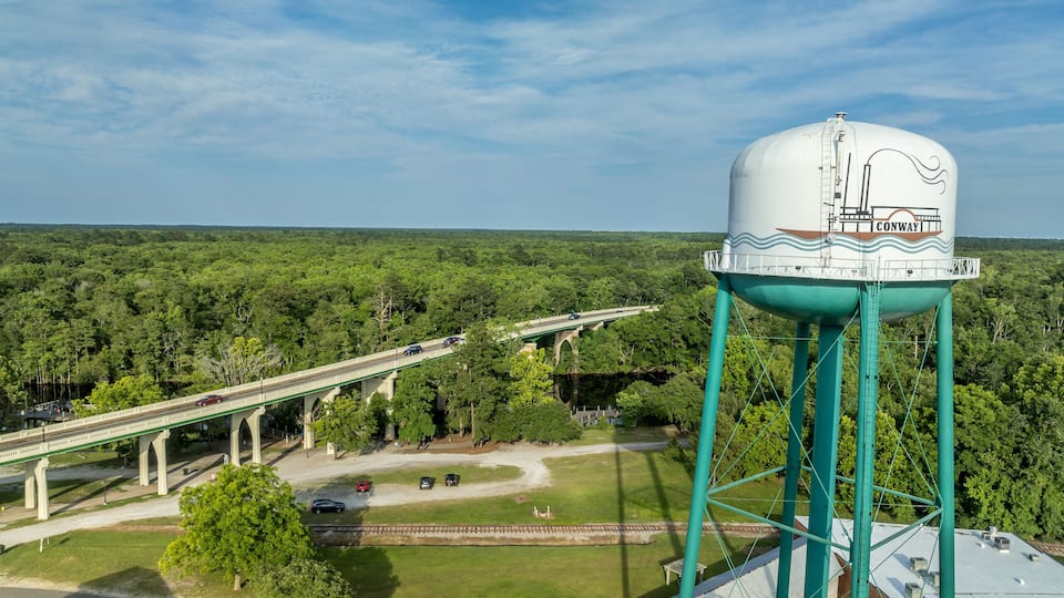 A water tower with 'CONWAY' written on it, a highway bridge, and a forested landscape.