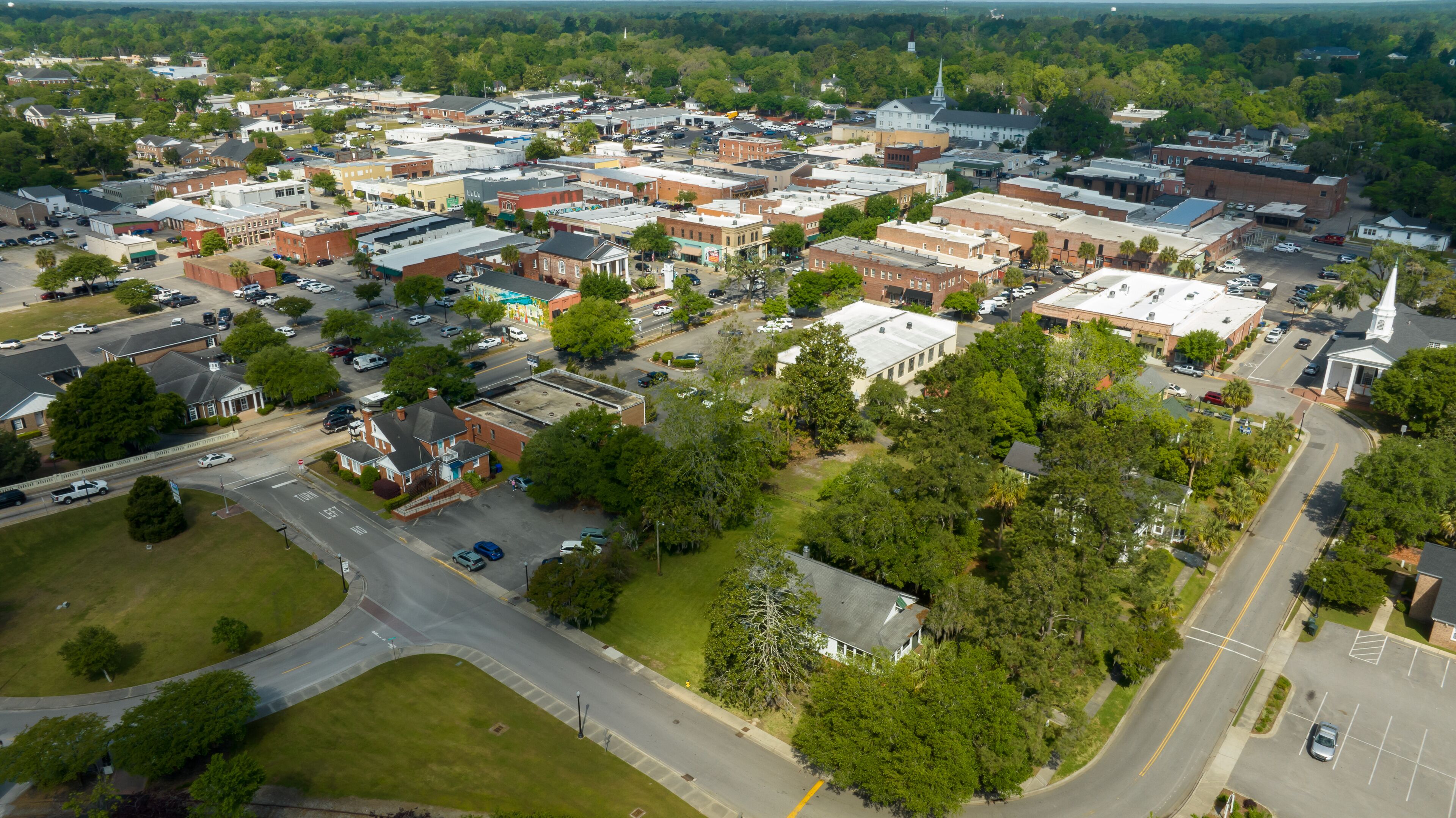 An aerial view of a town with a mix of commercial and residential buildings, a church steeple, and a large green park area.