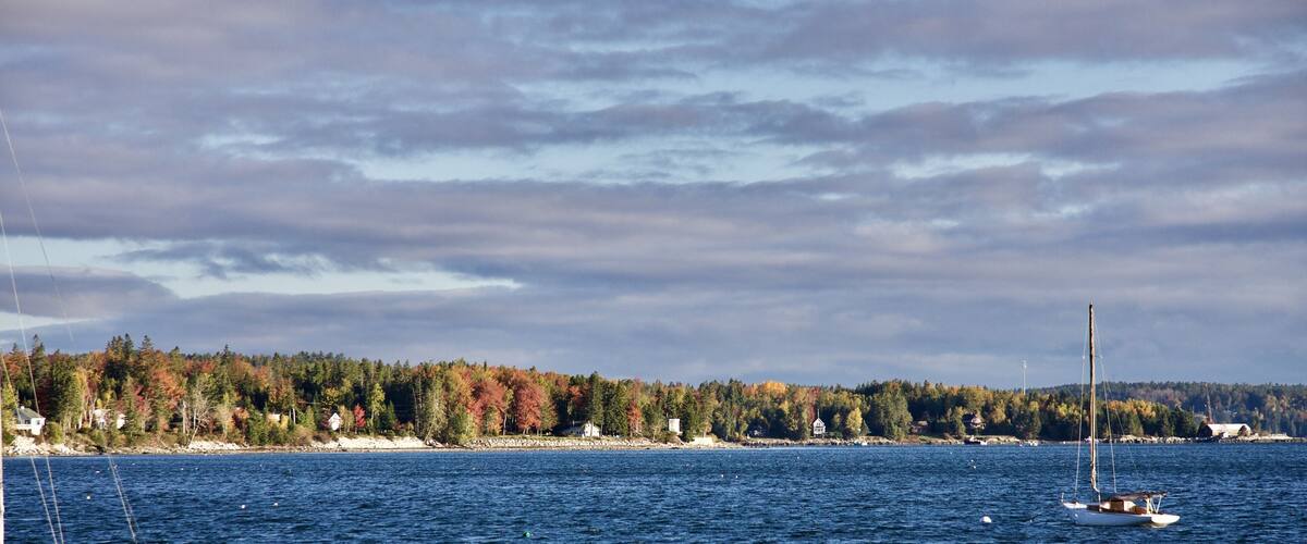 A calm sea with a sailboat, a forested shoreline, and a cloudy sky.