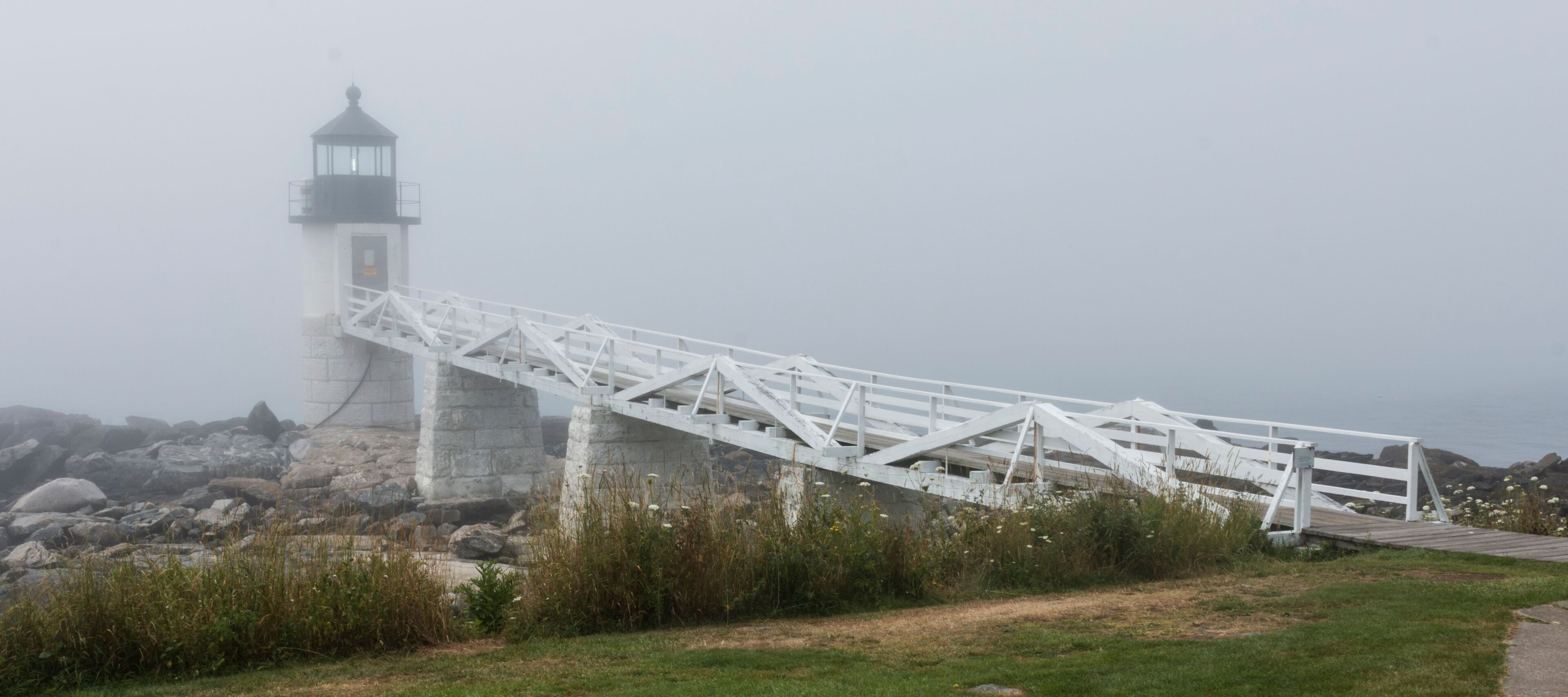 A white metal bridge leading to a lighthouse on a rocky shore.