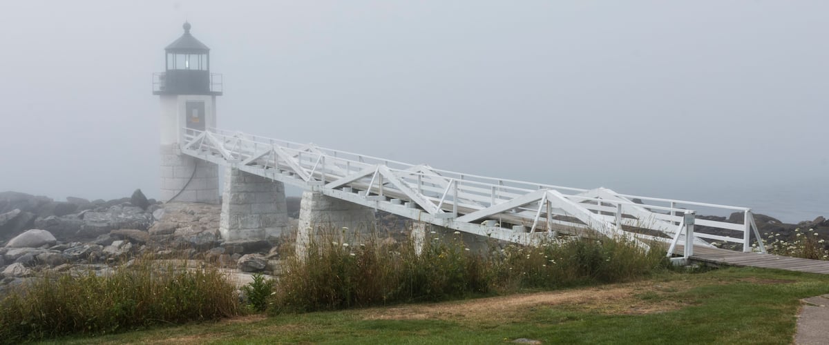 A white metal bridge leading to a lighthouse on a rocky shore.