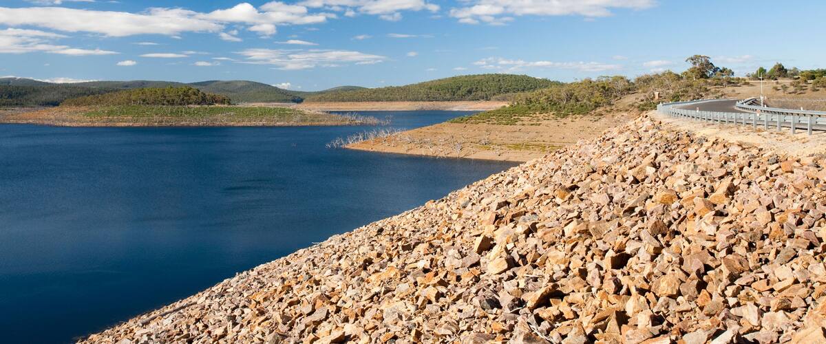 A rocky shoreline with a bridge over a calm lake, surrounded by hills and sparse vegetation.