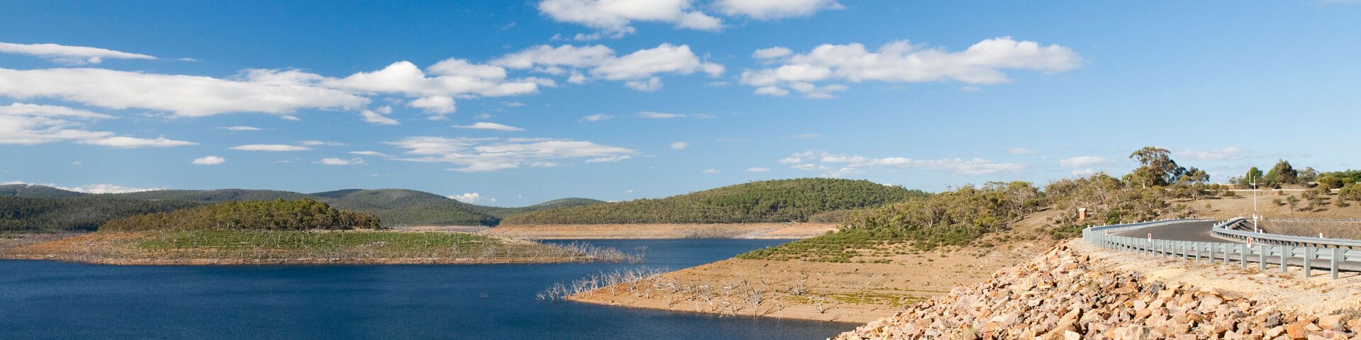 A rocky shoreline with a bridge over a calm lake, surrounded by hills and sparse vegetation.