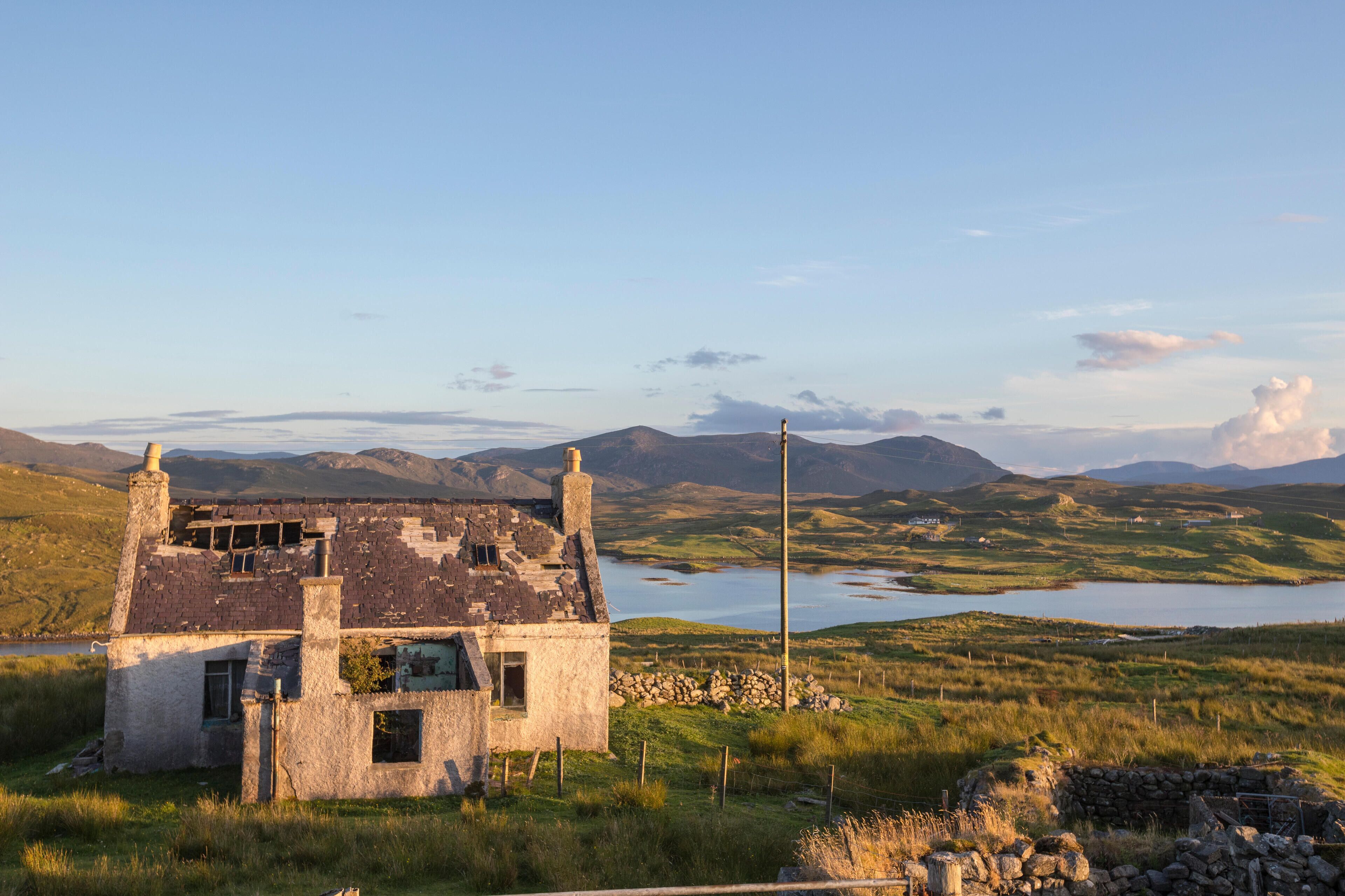 A dilapidated stone house with a chimney, surrounded by a grassy landscape and a lake, with mountains in the background.