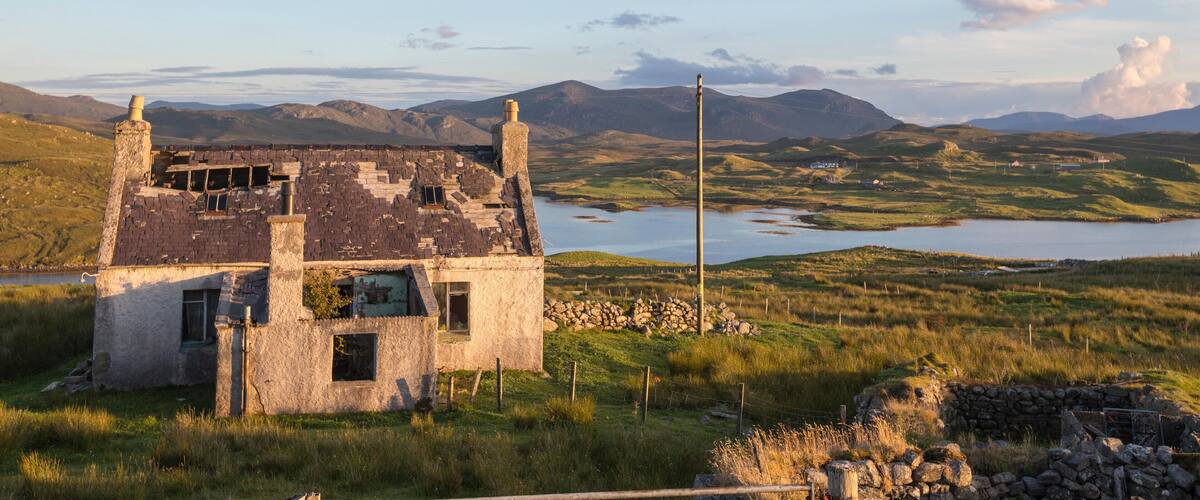 A dilapidated stone house with a chimney, surrounded by a grassy landscape and a lake, with mountains in the background.