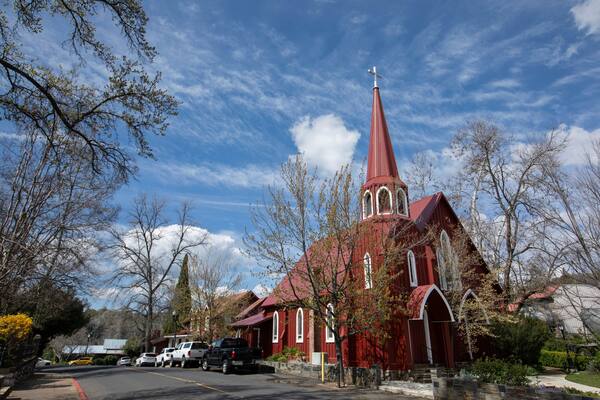 Une église rouge, dotée d’un clocher élevé et surmontée d’une croix, entourée d’arbres et de voitures garées.