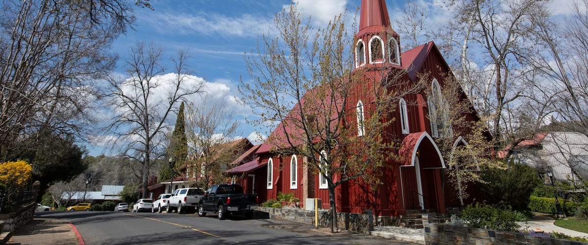 A red church with a tall steeple and a cross on top, surrounded by trees and parked cars.