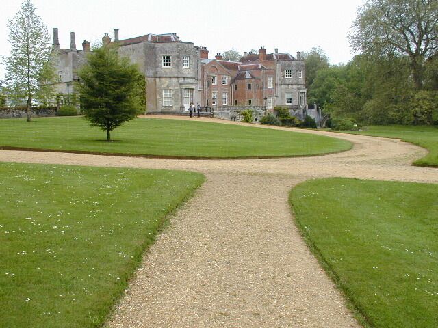 A gravel path leading to a large, historic building surrounded by well-maintained lawns and trees.