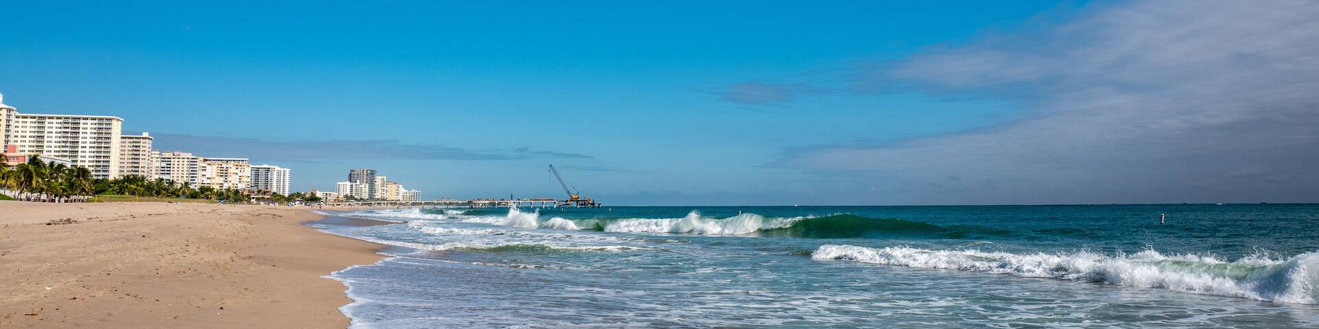A beach with waves, a pier, and buildings in the background.