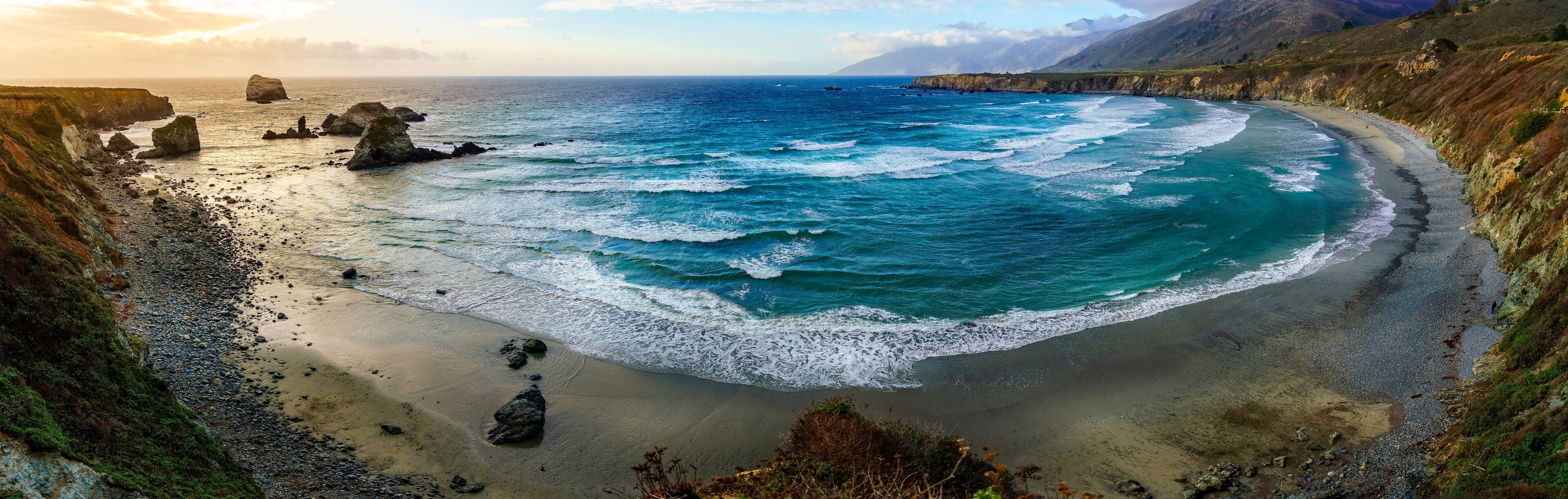 A coastal landscape with a sandy beach, rocky outcrops, and a clear view of the ocean.