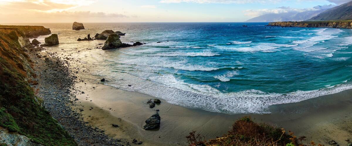 A coastal landscape with a sandy beach, rocky outcrops, and a clear view of the ocean.