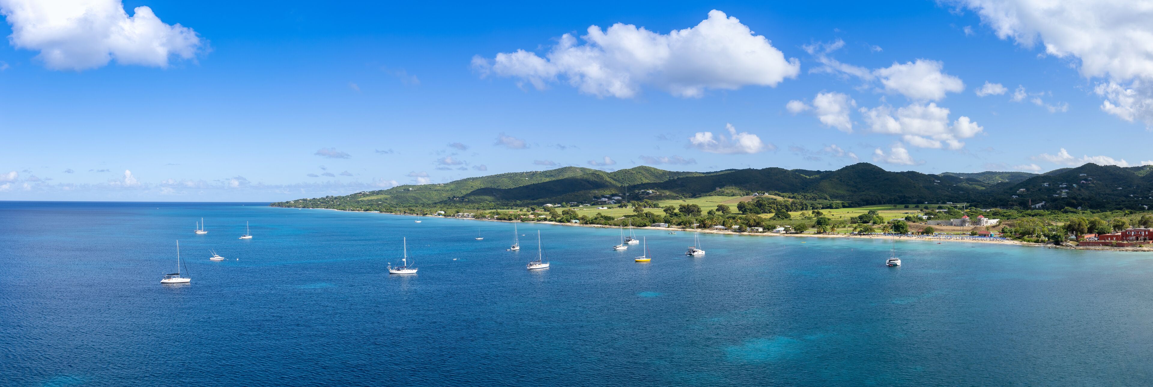 A coastal landscape with clear blue waters, several sailboats, and green hills in the background.
