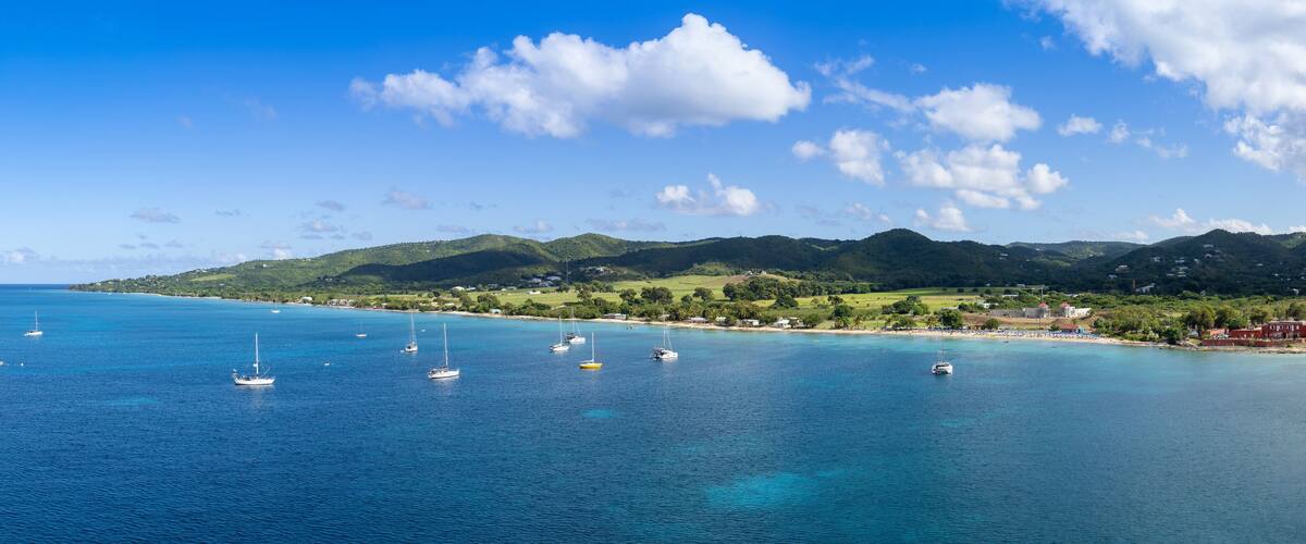 A coastal landscape with clear blue waters, several sailboats, and green hills in the background.