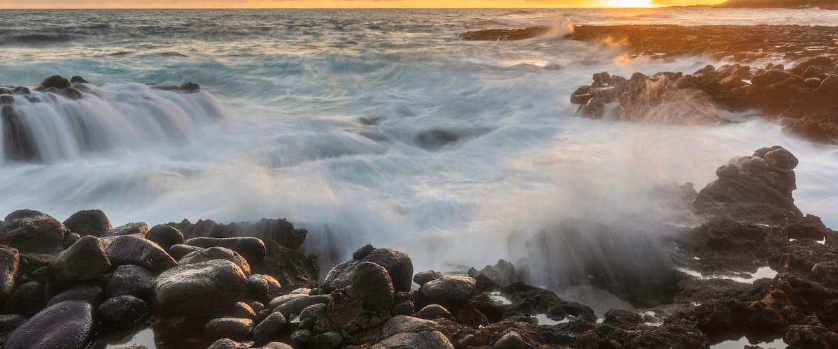 A rocky shoreline with waves crashing against the rocks during sunset.