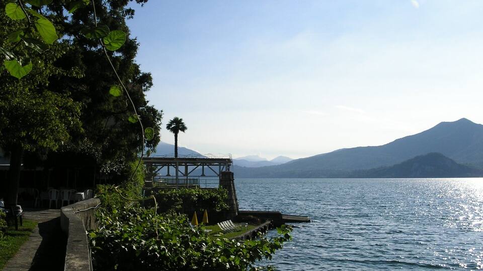 A lakeside walkway with a palm tree, a dock, and mountains in the background.
