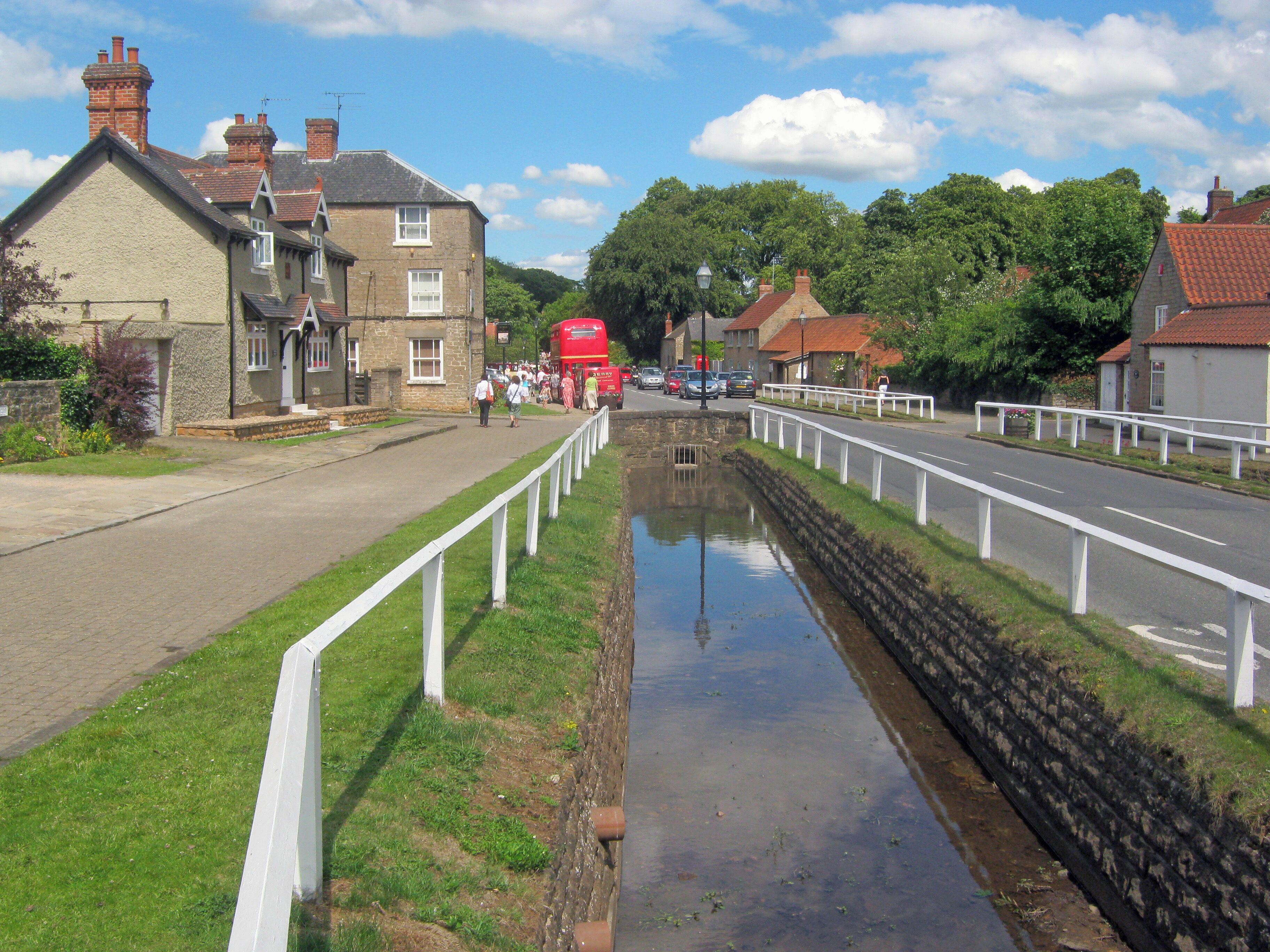 A canal with a white fence, a red double-decker bus, and houses with red roofs.