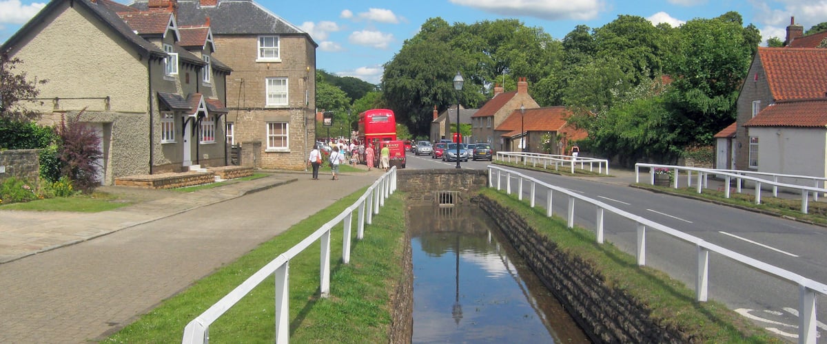 A canal with a white fence, a red double-decker bus, and houses with red roofs.