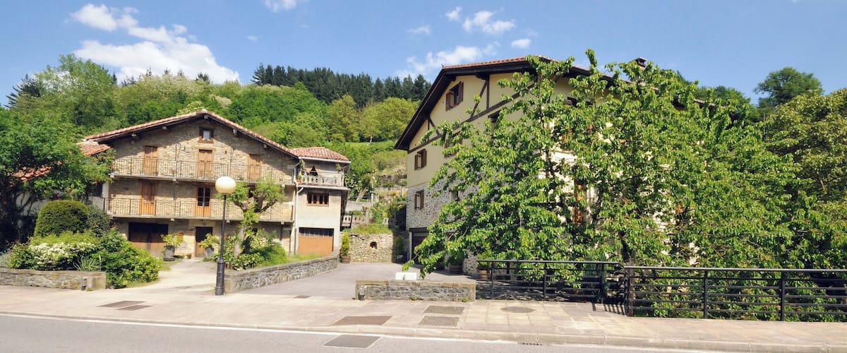 A rural street with stone buildings, a paved road, and a hillside with trees.