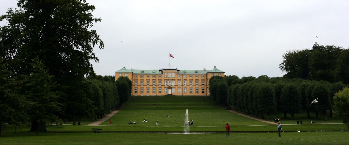 A large, historic building with a green roof, surrounded by a well-manicured lawn and a fountain.