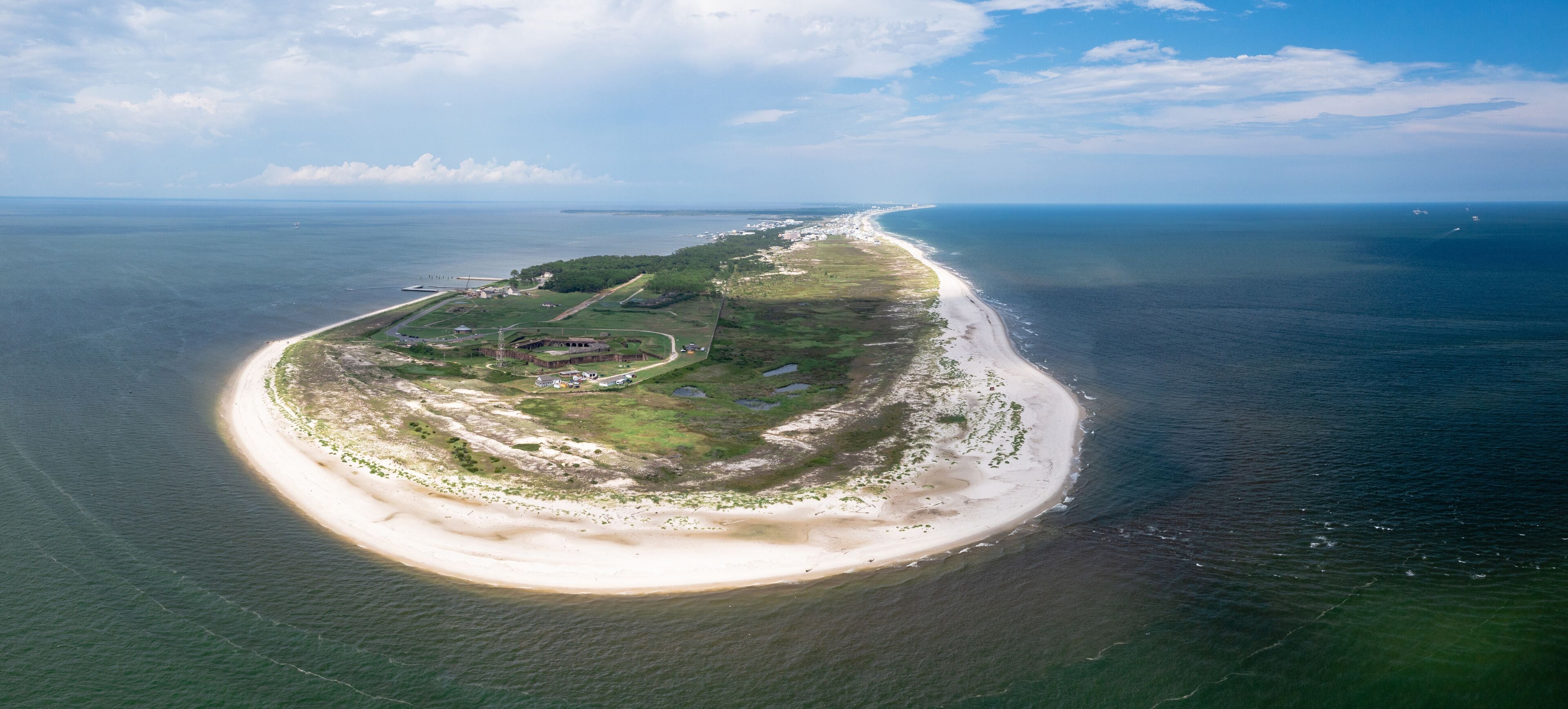 An aerial view of a coastal island with a sandy beach, green vegetation, and a clear view of the ocean.