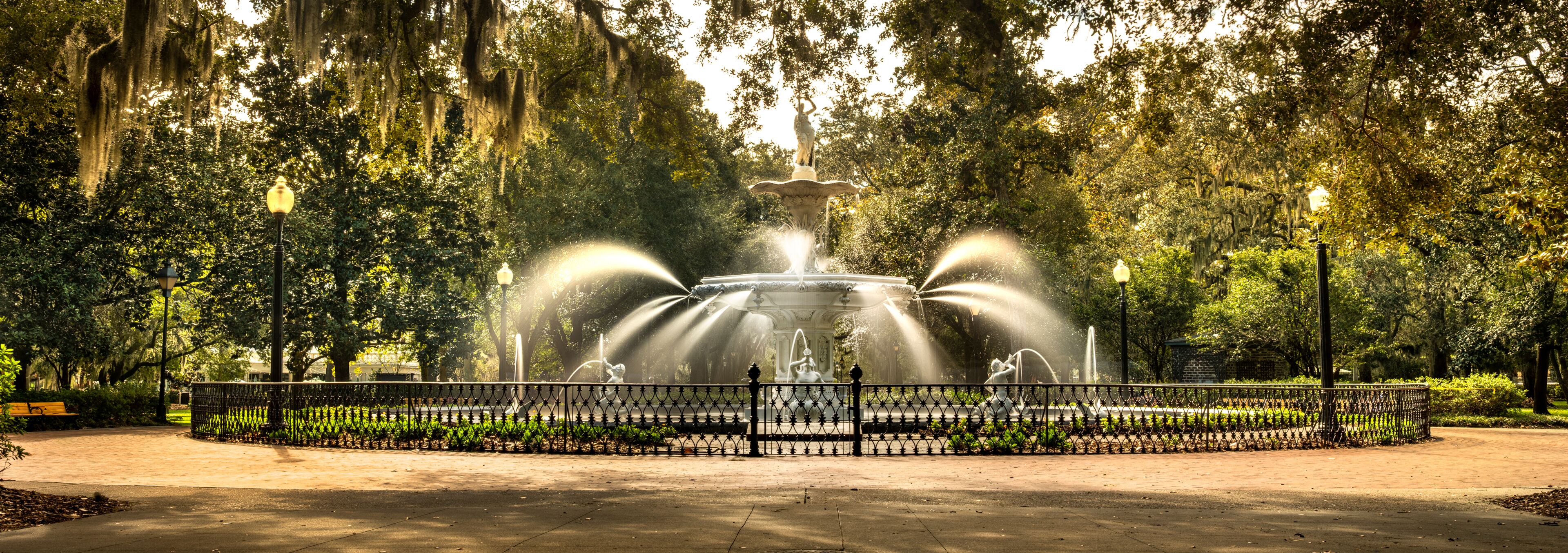 A fountain with water jets surrounded by a fence, set in a park with trees and a bench.