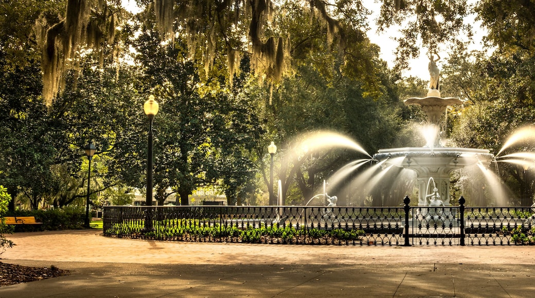 A fountain with water jets surrounded by a fence, set in a park with trees and a bench.