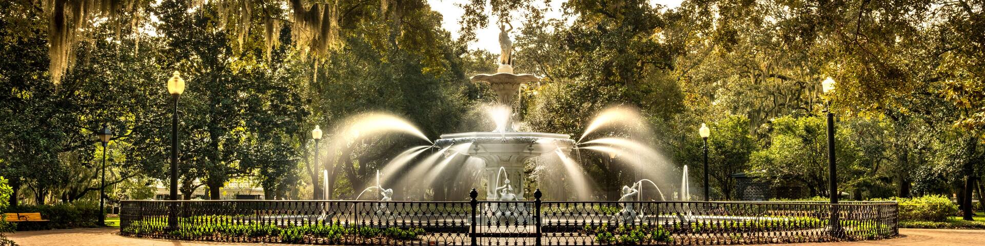 A fountain with water jets surrounded by a fence, set in a park with trees and a bench.