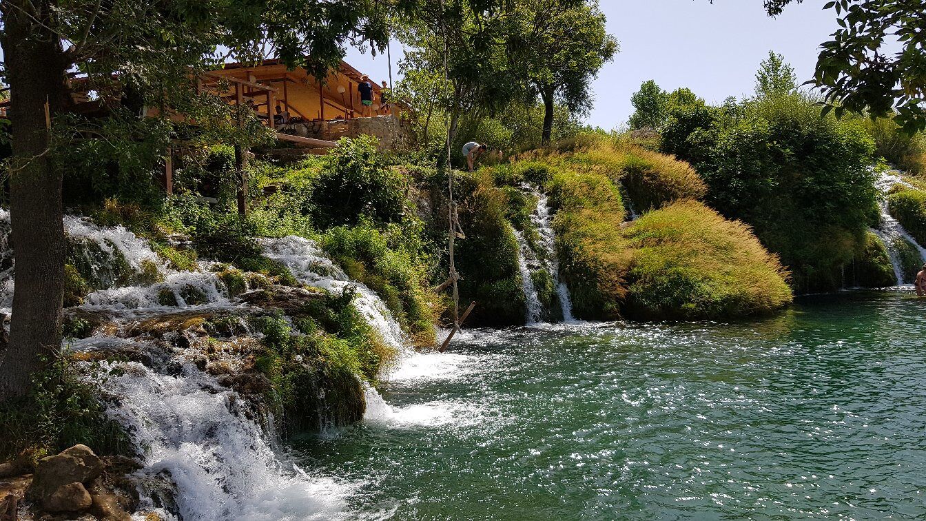 A waterfall cascading over rocks into a river surrounded by lush greenery and a wooden structure in the background.
