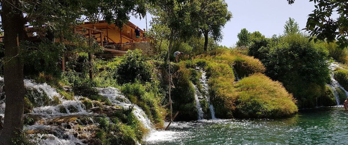 A waterfall cascading over rocks into a river surrounded by lush greenery and a wooden structure in the background.