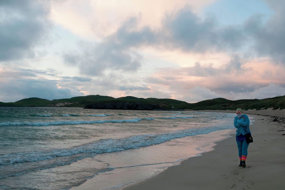 A beach with waves, a cloudy sky, and green hills in the background.