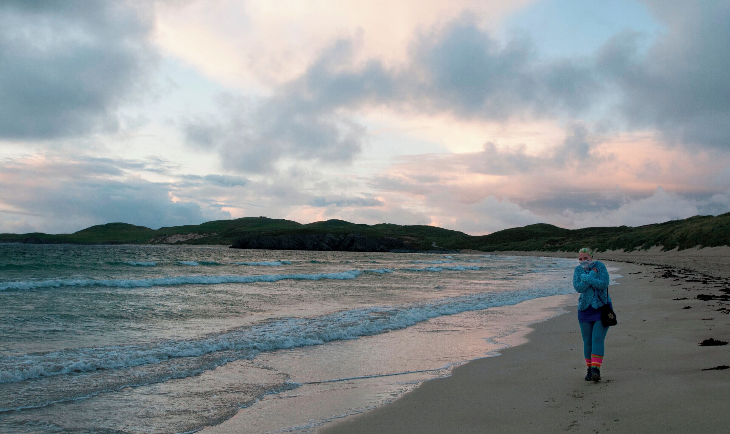 A beach with waves, a cloudy sky, and green hills in the background.