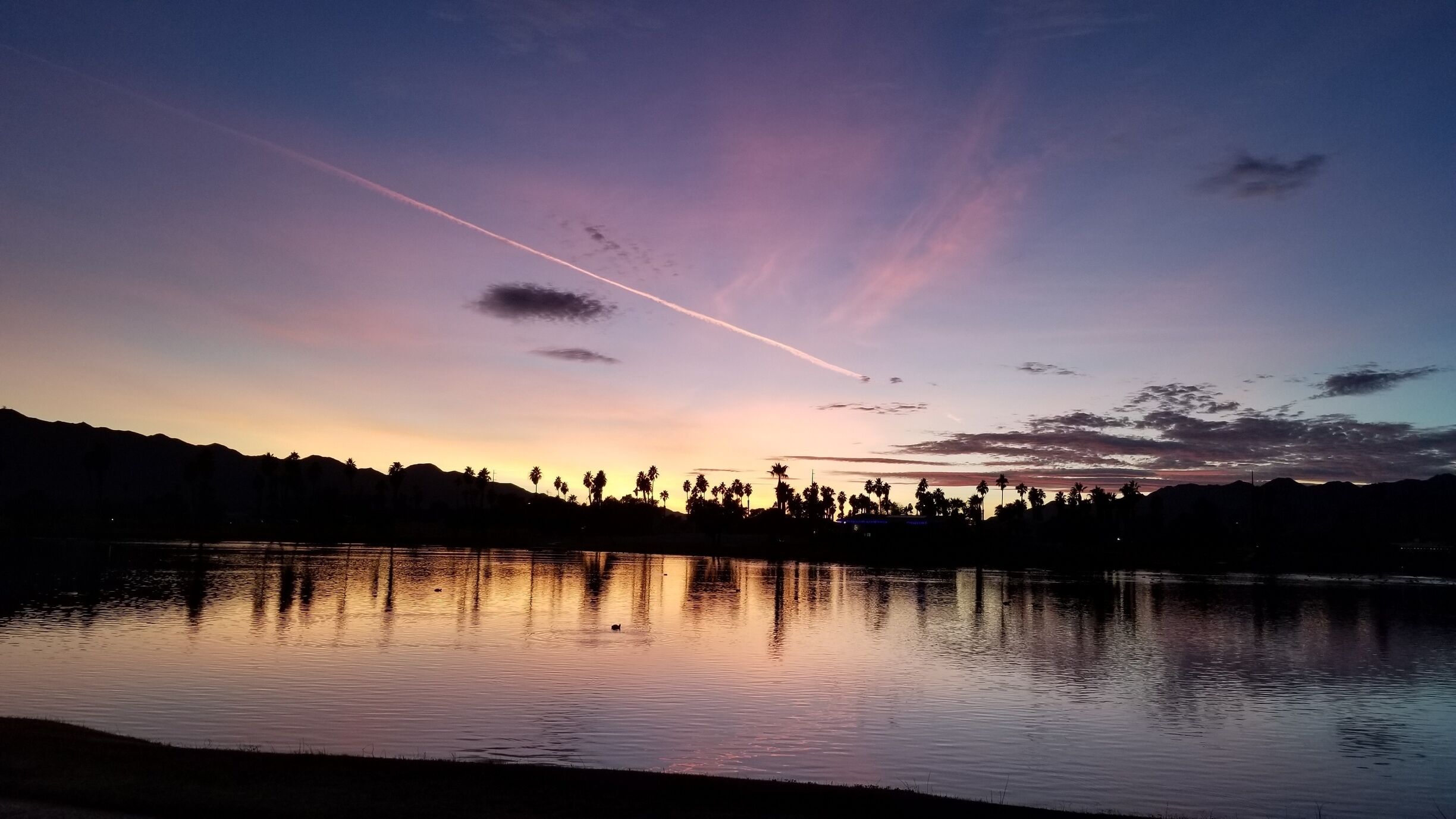 A sunset over a calm lake with silhouettes of palm trees and mountains in the background.