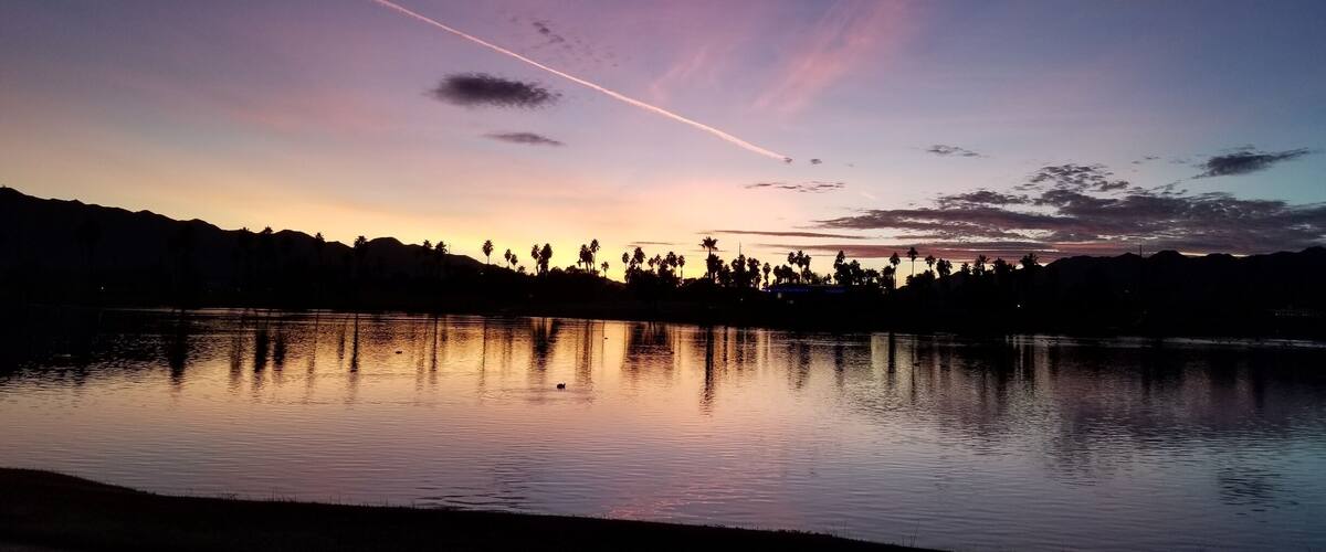 A sunset over a calm lake with silhouettes of palm trees and mountains in the background.