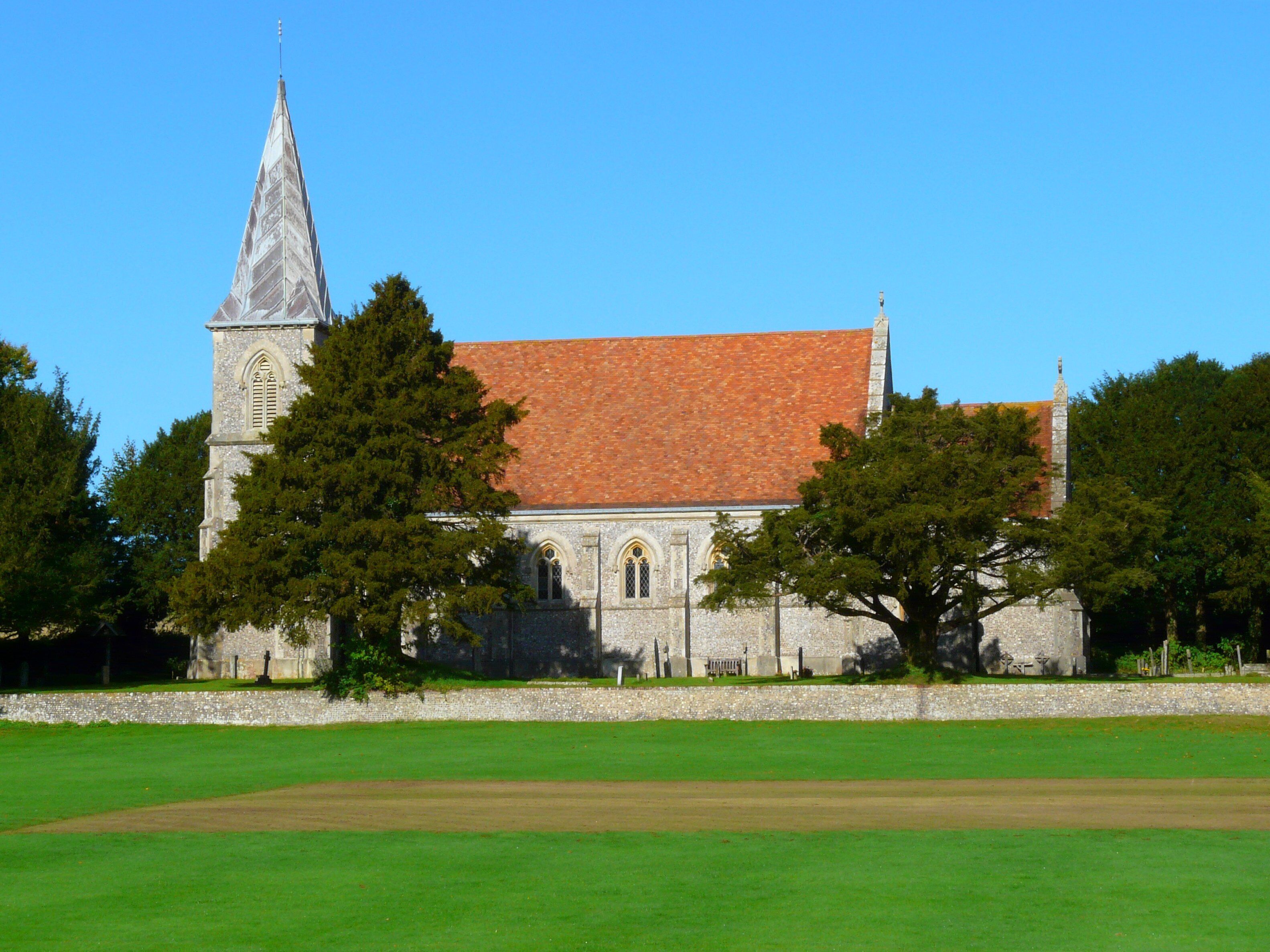 A church with a tall steeple and a red-tiled roof, surrounded by green trees and a grassy field.