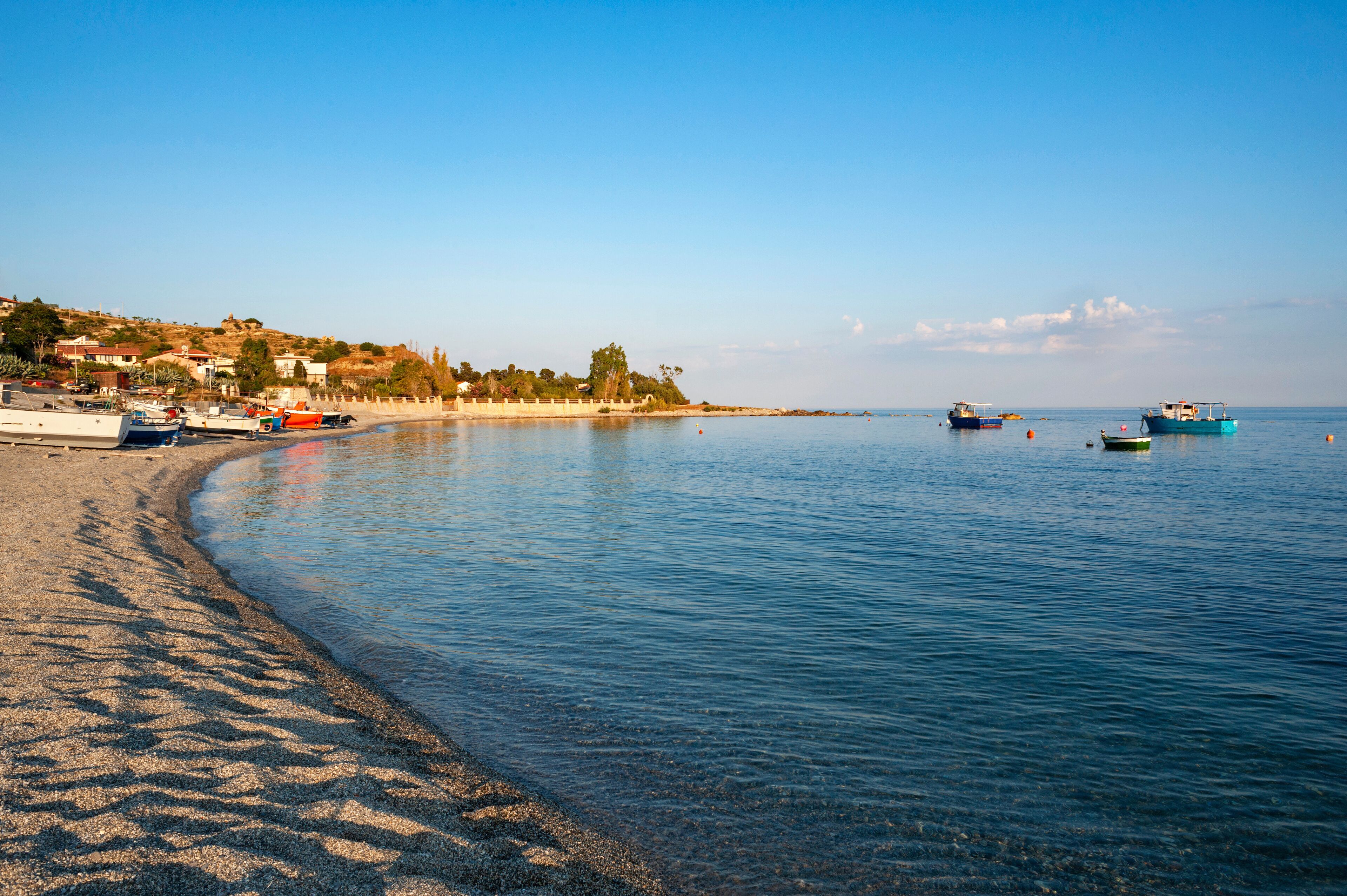 A beach with boats, clear water, and a hilly landscape in the background.
