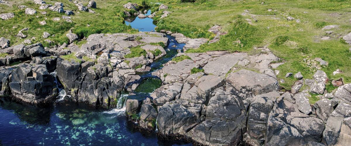 A rocky landscape with a small stream flowing through it, surrounded by green hills and a distant town.
