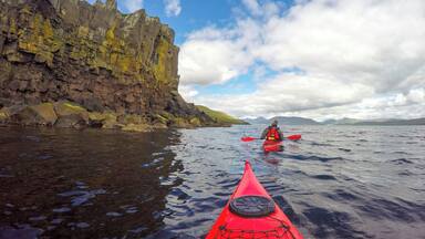 A kayaker in a red kayak on a calm sea with rocky cliffs in the background.