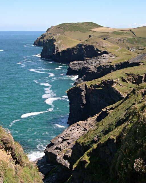A coastal cliff with waves crashing against it, a grassy hill in the background, and a clear sky.