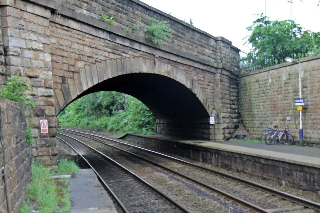 Uma ponte ferroviária de pedra com trilhos abaixo, uma plataforma à direita e uma placa indicando 'Greenfield' ao fundo.