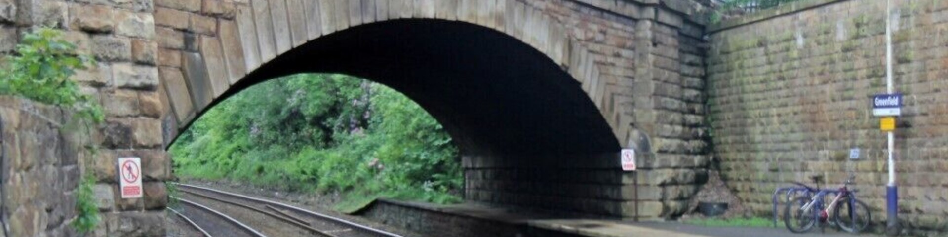 Uma ponte ferroviária de pedra com trilhos abaixo, uma plataforma à direita e uma placa indicando 'Greenfield' ao fundo.