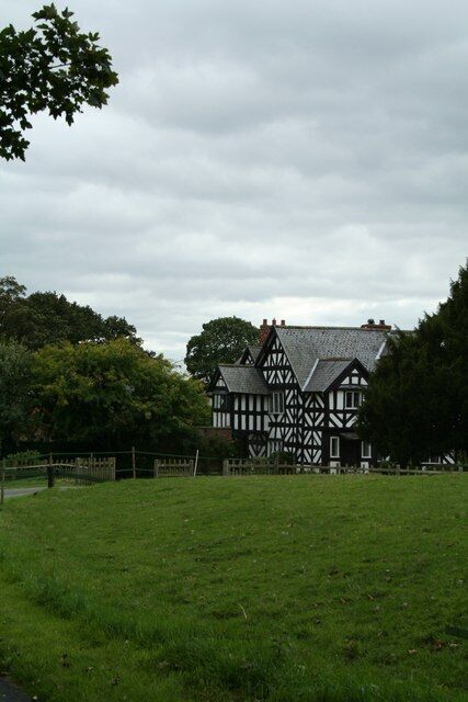 A Tudor-style house with a thatched roof, surrounded by a green field and trees.