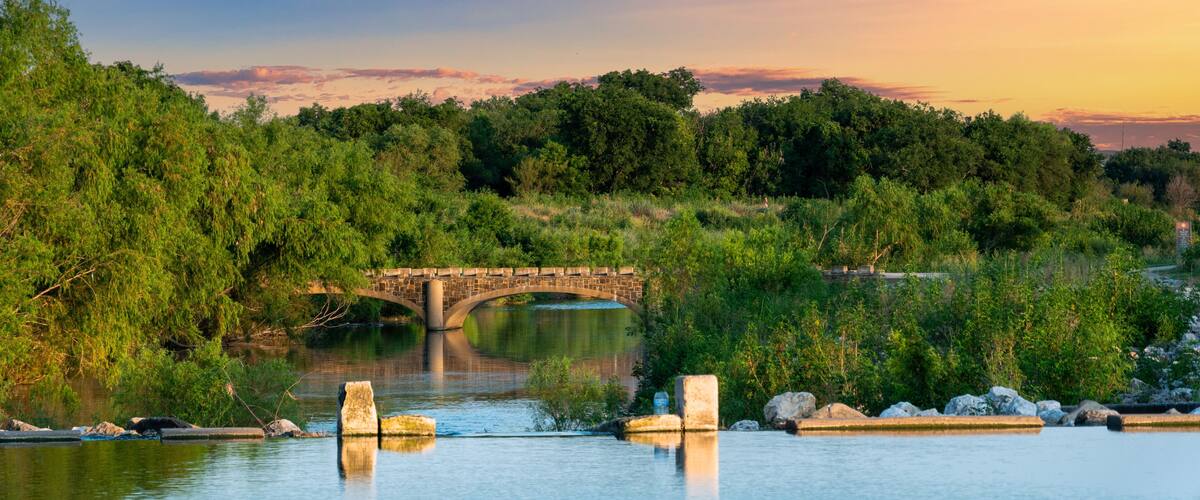 A stone bridge over a calm river, surrounded by lush greenery and trees.
