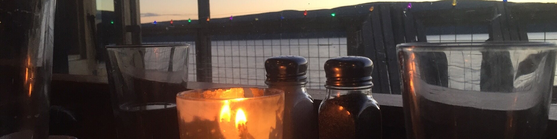 Mesa com copos e saleiros perto de uma janela com vista para a água ao pôr do sol.