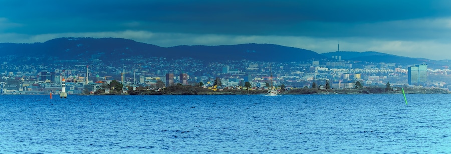 Un paisaje costero con un faro destacado, una zona urbana densa y una cadena montañosa al fondo.