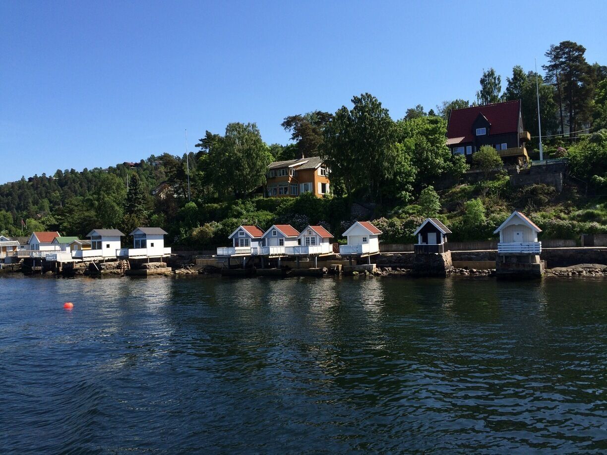 A waterfront with several small houses on stilts, a larger house on a hill, and a clear blue sky.