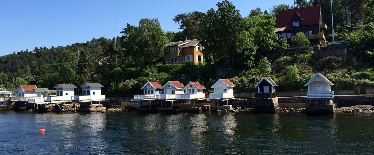 A waterfront with several small houses on stilts, a larger house on a hill, and a clear blue sky.