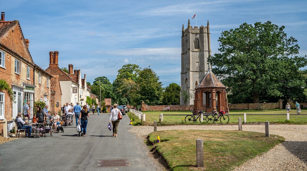 A town square with a church tower, a red-brick building with a sign, and people walking and sitting on benches.
