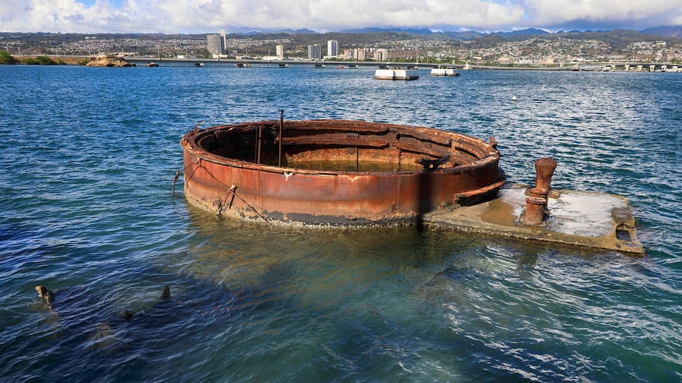 A rusted, circular metal structure in the water with a cityscape in the background.