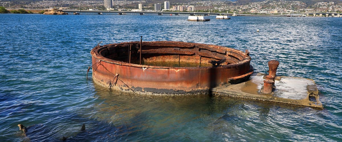 A rusted, circular metal structure in the water with a cityscape in the background.
