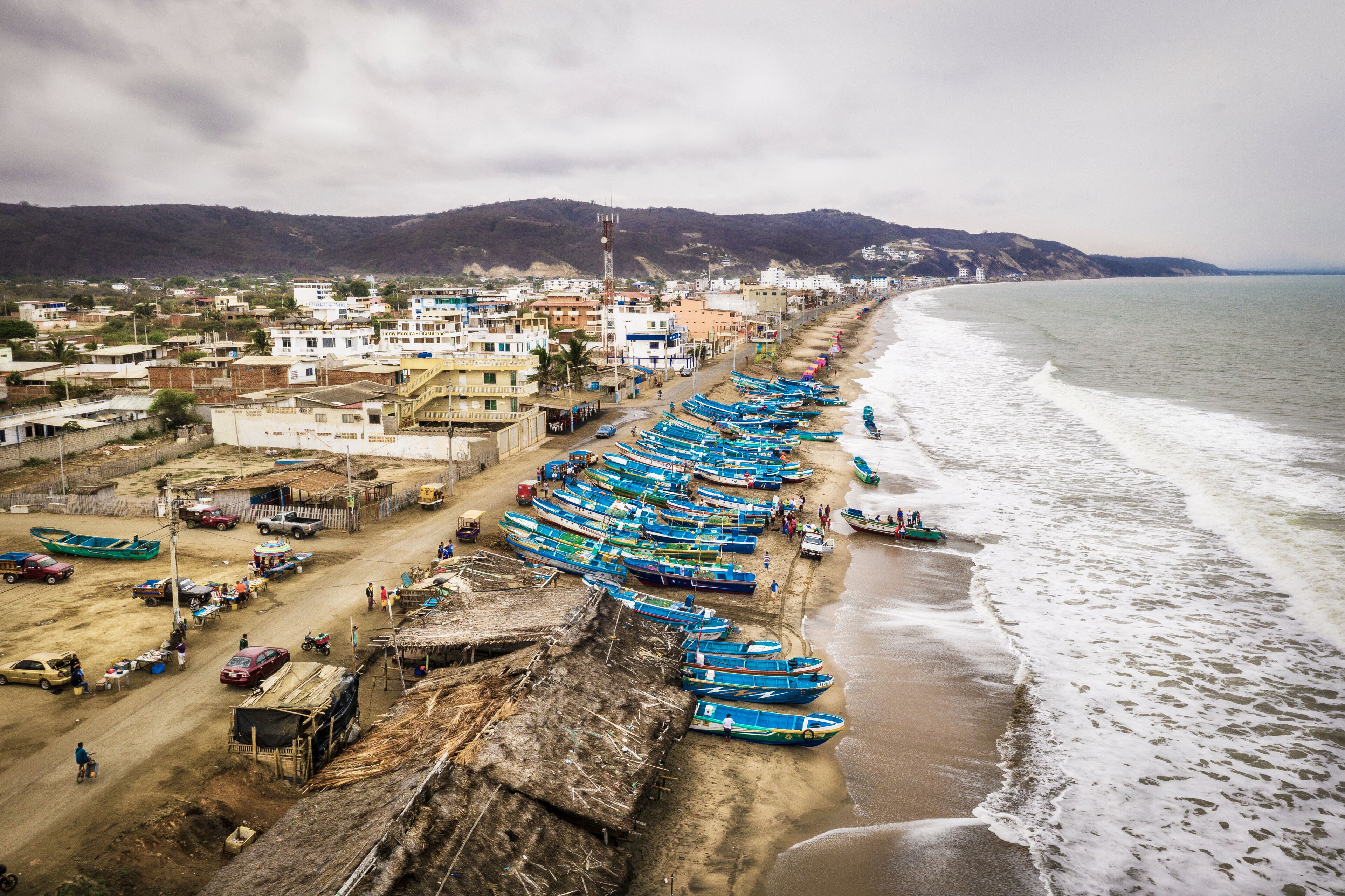 A coastal town with a long row of boats lined up along the beach, a dirt road, and buildings in the background.